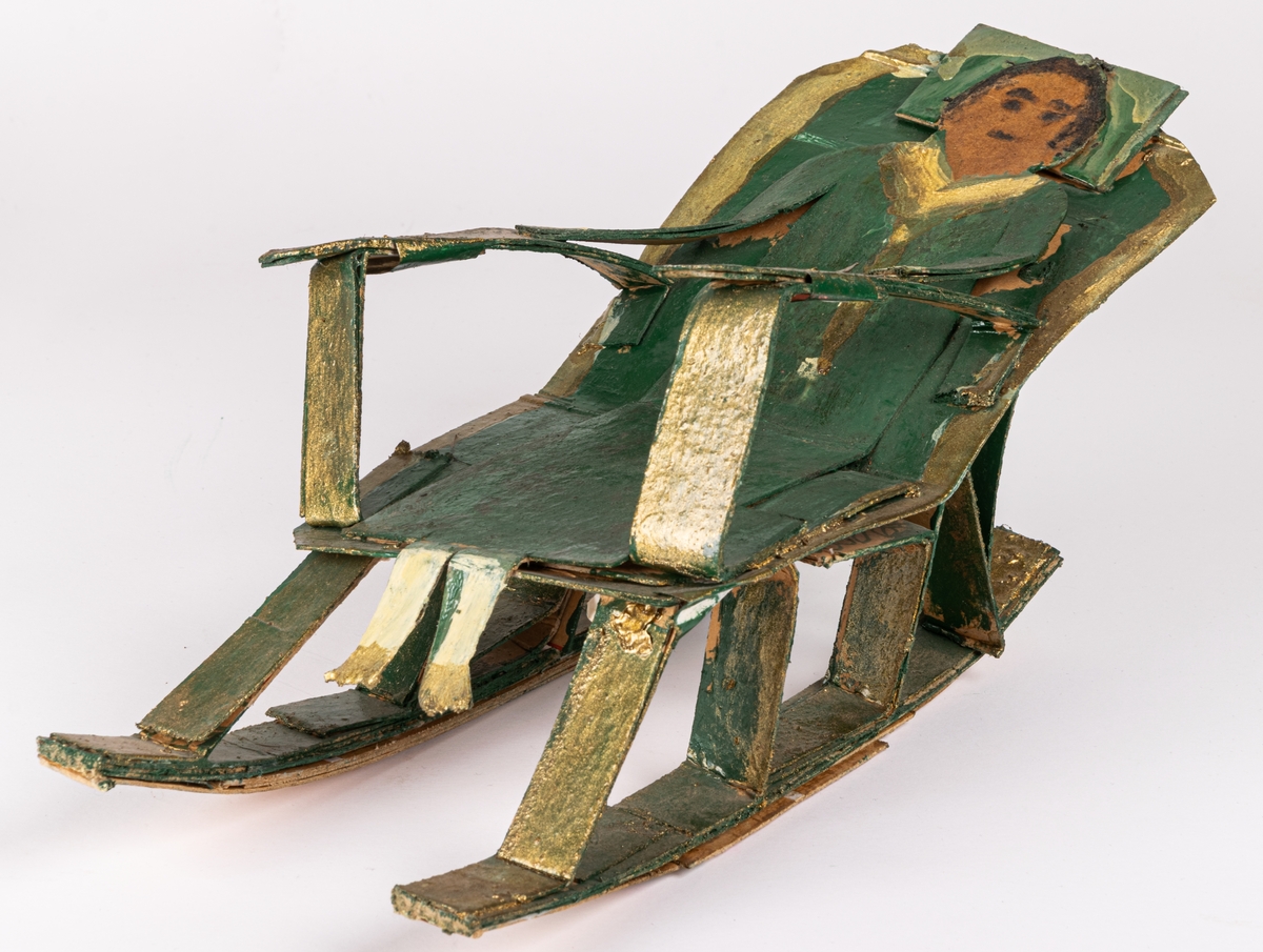 Skulptur av papp, oljefärg och troligen lackfärg. Föreställer grönklädd kvinna i gungstol. Målad i grönt och guld.