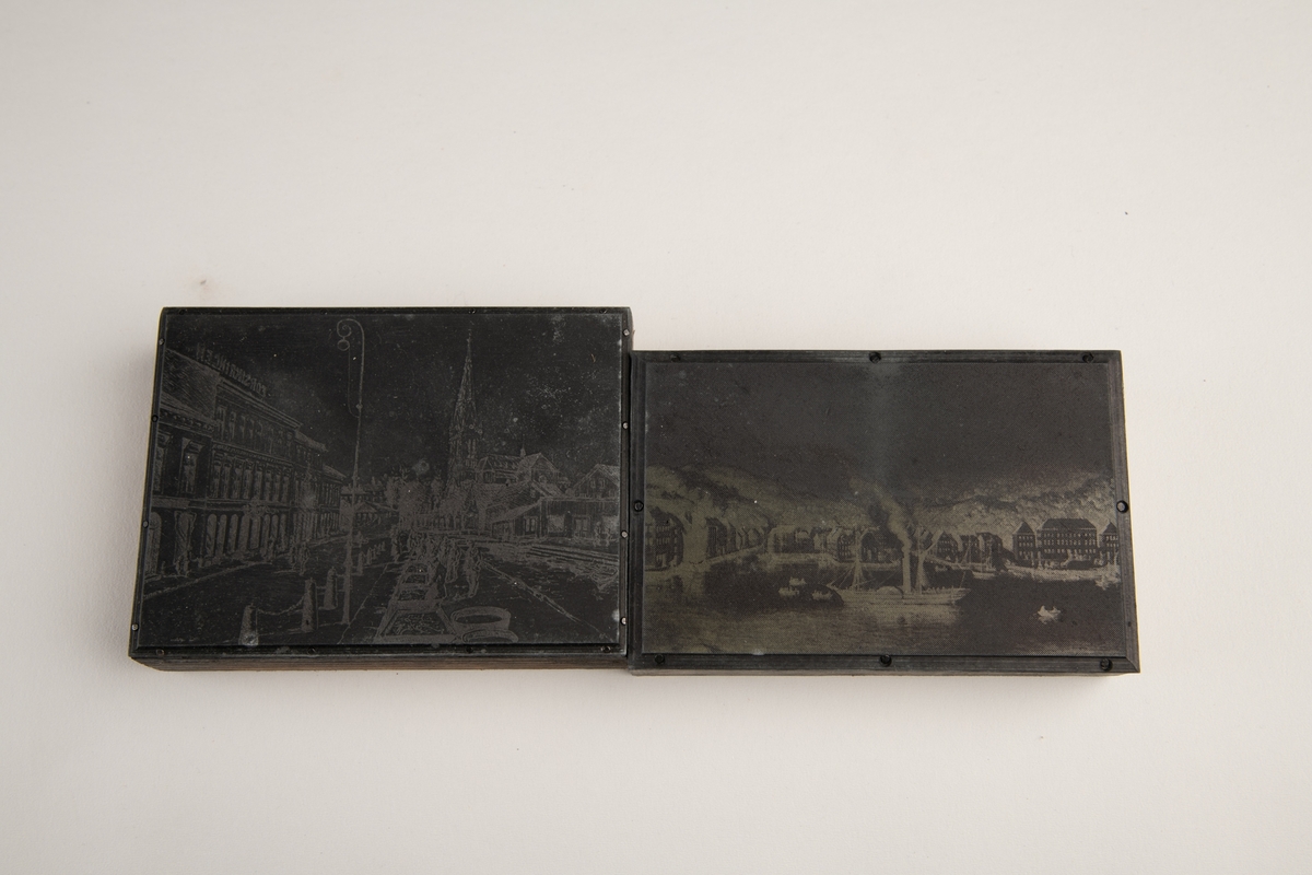 To klisjeer i en konvolutt. Kovolutten har påskrift i svart og blå tusj samt bå kulepenn. Den ene klisjeen er et bilde av en dampbåt på sjøen omringet av småbåter og med havnebygg i bakgrunnen. Delvis utydelig skrift på den ene siden. På motsatt side av trykkeplaten er det festet papir. Den andre klisjeen har motiv av et bylandskap. Folk i gatne, en lyktestolpe i forgrunnen og en kirke i bakgrunnen. En papirlapp er festet på treklossen på andre siden av trykkplaten.