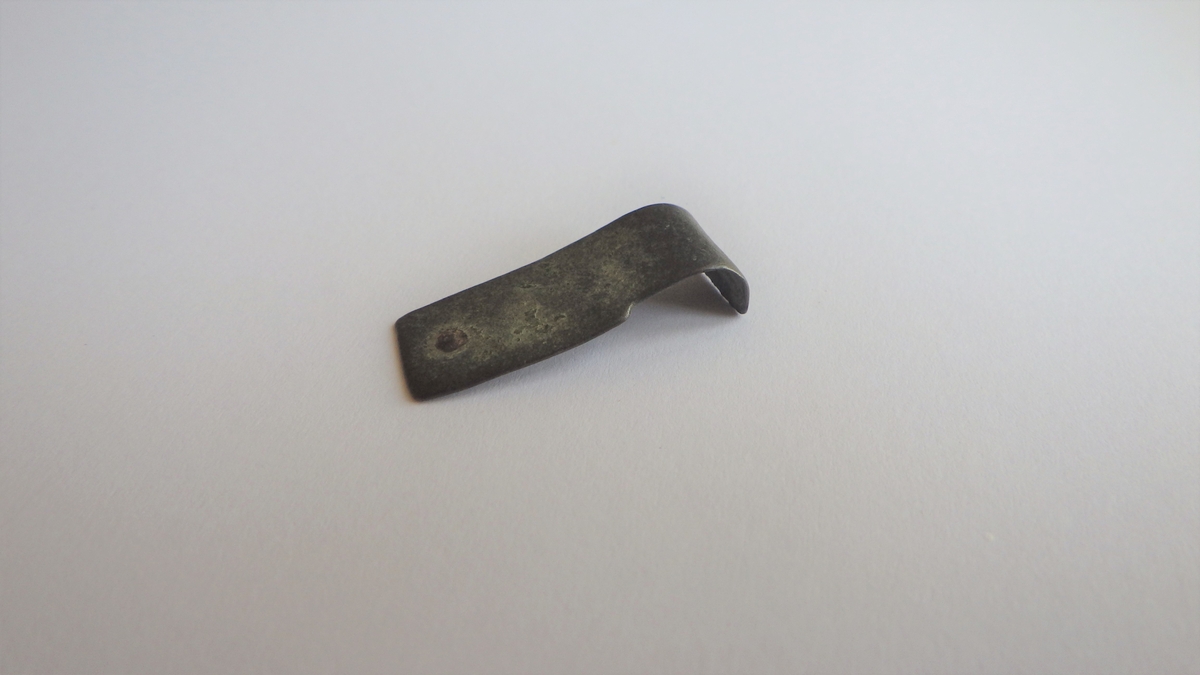 MALV: L. 25 MM  B. 10 MM, BÅNDFORMET UTEN DEKOR. REST AV EN NAGLE I ENE ENDEN. DEN ANDRE ENDEN ER OMBØYET. 