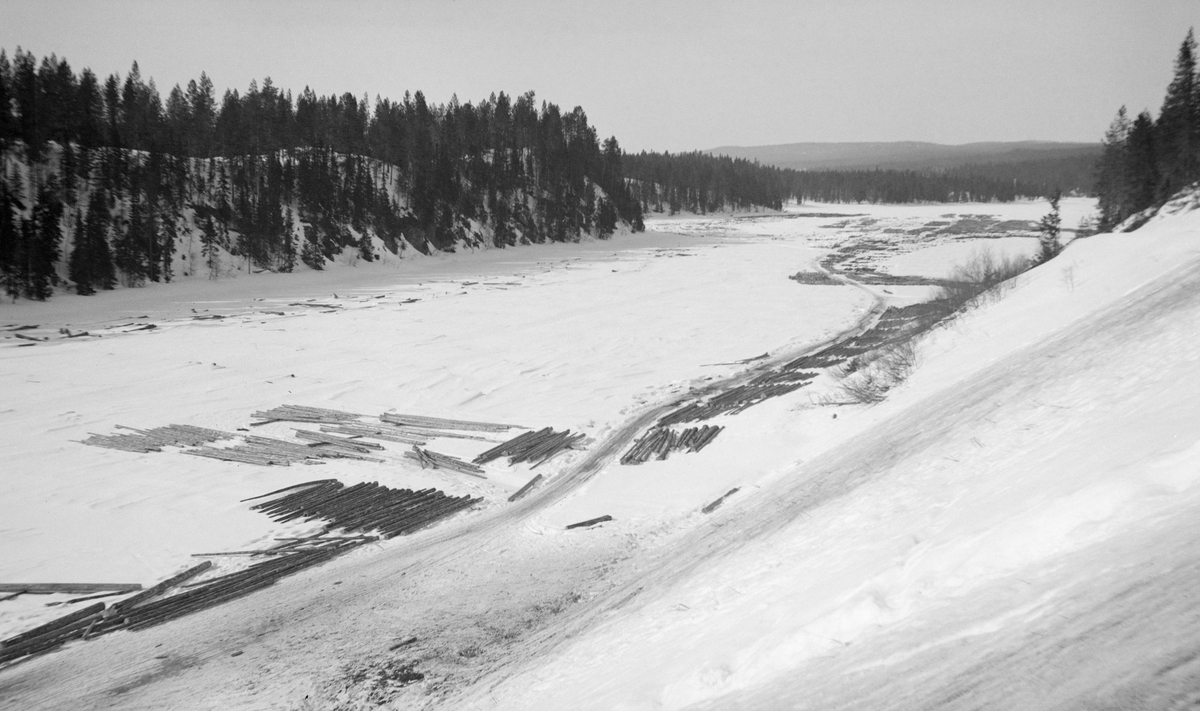 Fra Vesle-Osen i sørenden av Osensjøen i Trysil.  Fotografiet er tatt vinteren 1934.  Bildet viser hvordan tømmeret ble utlagt på den islagte og snødekte sjøen i påvente av våren og fløtingssesongen.  Fotografen har stått i en skråning med kameraet vendt ned mot isflata.  Kjørevegen og det sist avlessete tømmeret framtrer klart på bildet, men i det skrå sollyset aner vi at det også ligger en god del tømmerstokker skjult under snøen. 