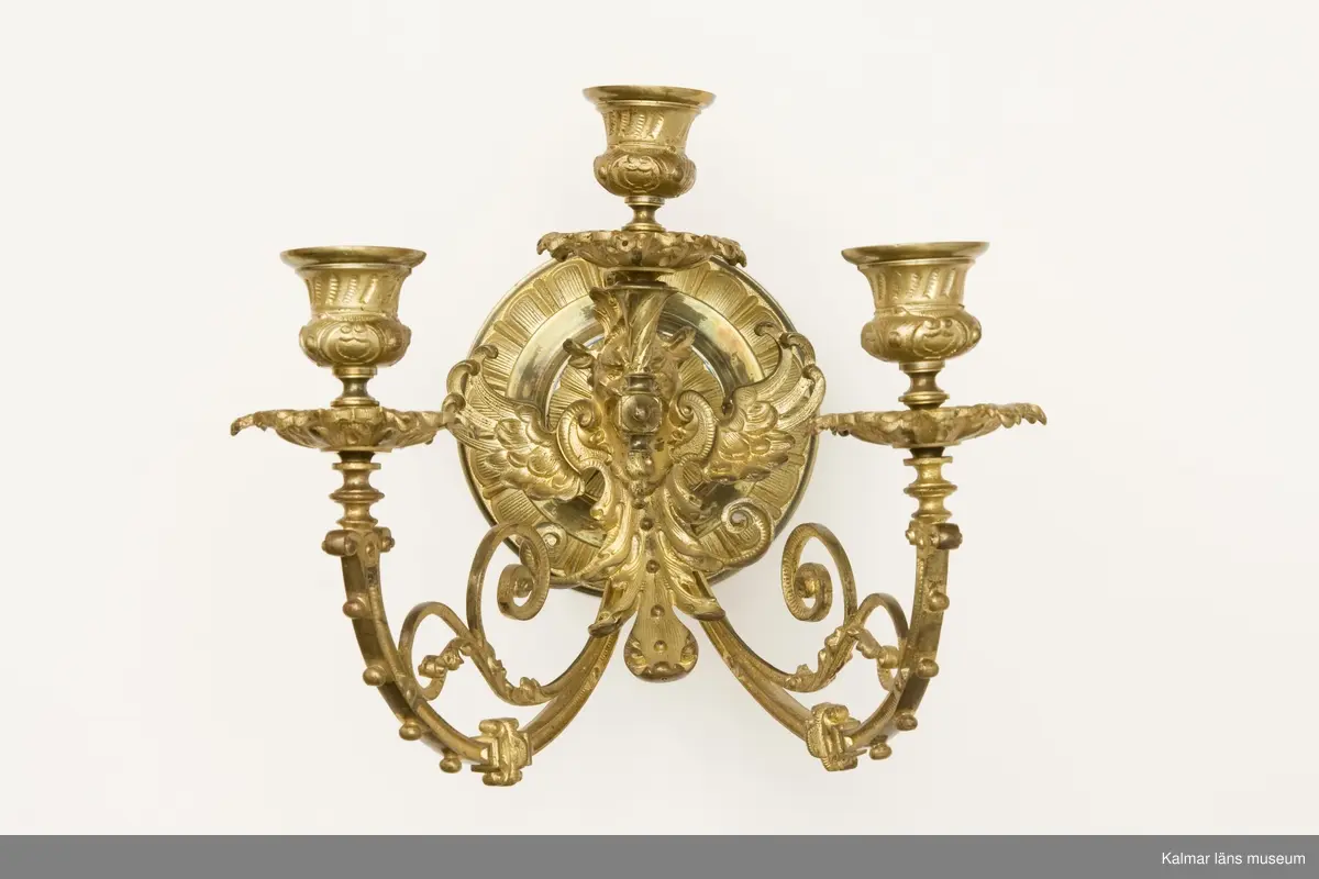 KLM 31995:1-2 Ljusstakar, lampetter, ett par, mässing. Gjutna av mässing. Tre ljushållare på respektive lampett. Ornamentik i form av bevingat djurhuvud, blad och växtslingor.