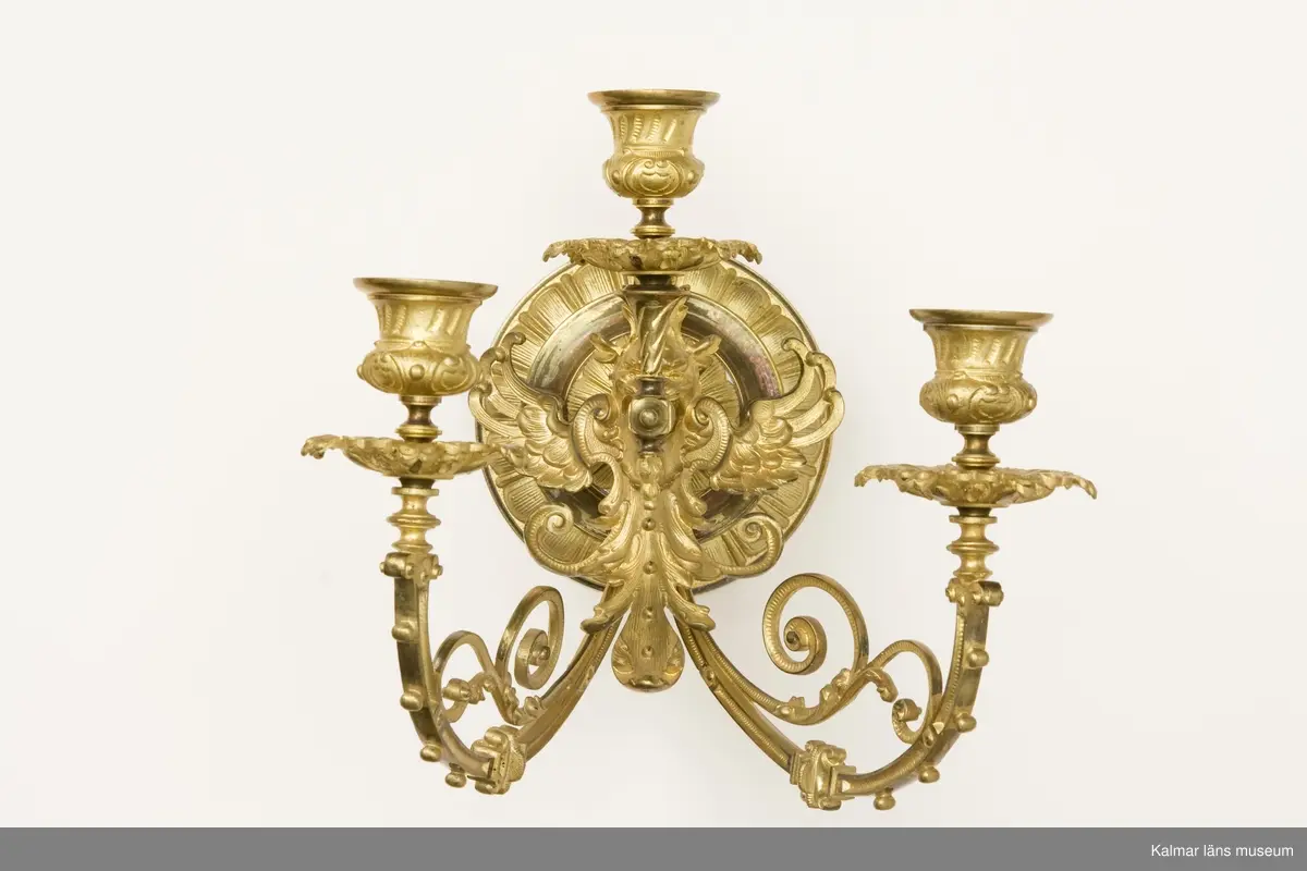 KLM 31995:1-2 Ljusstakar, lampetter, ett par, mässing. Gjutna av mässing. Tre ljushållare på respektive lampett. Ornamentik i form av bevingat djurhuvud, blad och växtslingor.