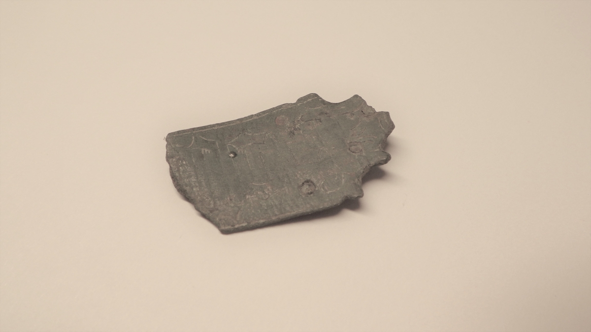 MALV: L. 36 MM B. 22 MM, TRAPESFORMET, AVBRUKKET I BEGGE ENDER. I DEN ENEENDEN ER DEN KLIPPET TIL I ET ORNAMENT SOM ANTAGELIG HAR ENDT I EN KROKELLER ET ØRE. I DENNE ENDEN SITTER OGSÅ FIRE NAGLER. ET NAGLEHULL ERAVBRUKKET. DEKORERT MED BOKSTAVER I STREKORNAMENTIKK, INNRISSET. 