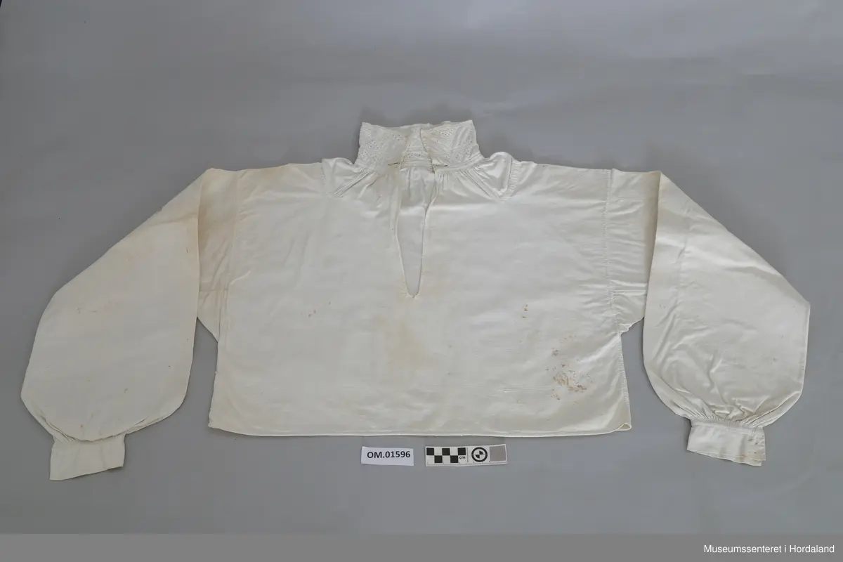 Kvit bomullskjorte med kvitsaumbroderi på krage. Kragen er høg og består av to border. To knapphol. Isett stoffbitar og rynking i hals. Splitt frå hals og ned halve skjorta. Kile under armane. Armane er rynka og har mansjettar utan broderi. Hekter. Ingen splitt i sidene. Handsauma. Eit hol i nakken. Flekkete.