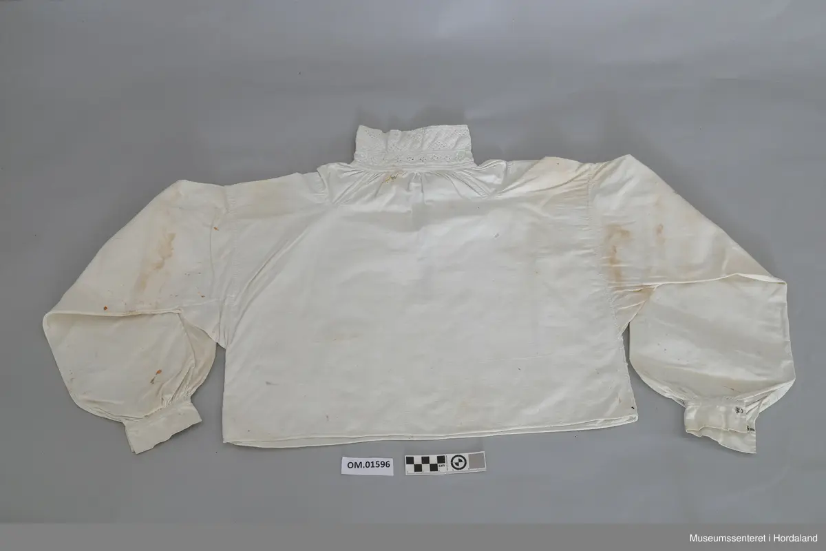 Kvit bomullskjorte med kvitsaumbroderi på krage. Kragen er høg og består av to border. To knapphol. Isett stoffbitar og rynking i hals. Splitt frå hals og ned halve skjorta. Kile under armane. Armane er rynka og har mansjettar utan broderi. Hekter. Ingen splitt i sidene. Handsauma. Eit hol i nakken. Flekkete.