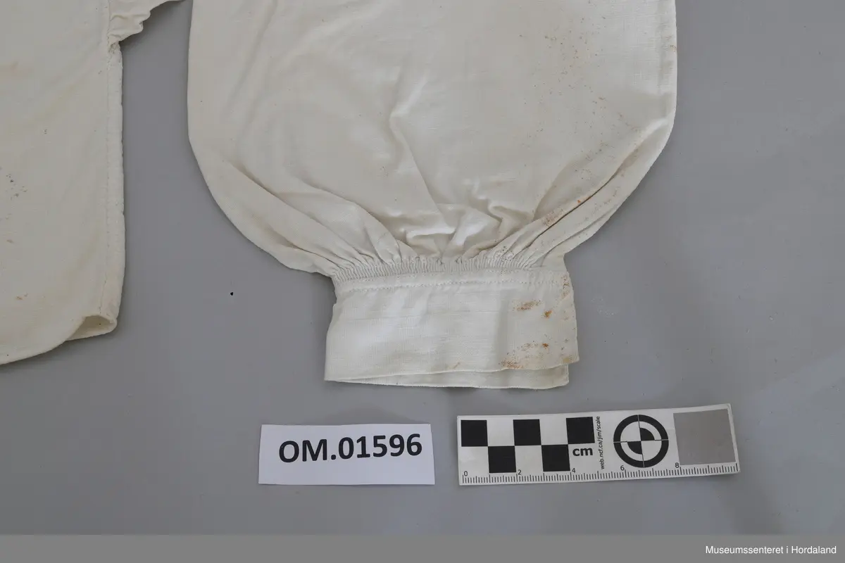 Kvit bomullskjorte med kvitsaumbroderi på krage. Kragen er høg og består av to border. To knapphol. Isett stoffbitar og rynking i hals. Splitt frå hals og ned halve skjorta. Kile under armane. Armane er rynka og har mansjettar utan broderi. Hekter. Ingen splitt i sidene. Handsauma. Eit hol i nakken. Flekkete.