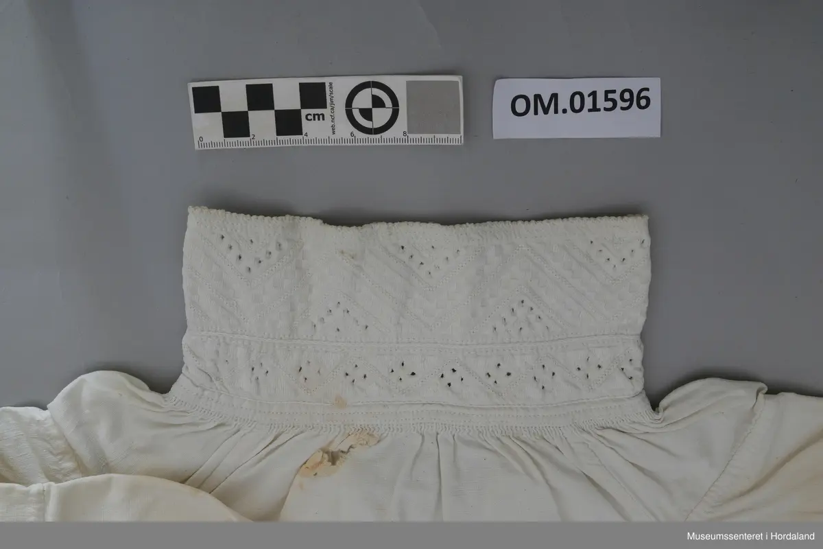 Kvit bomullskjorte med kvitsaumbroderi på krage. Kragen er høg og består av to border. To knapphol. Isett stoffbitar og rynking i hals. Splitt frå hals og ned halve skjorta. Kile under armane. Armane er rynka og har mansjettar utan broderi. Hekter. Ingen splitt i sidene. Handsauma. Eit hol i nakken. Flekkete.