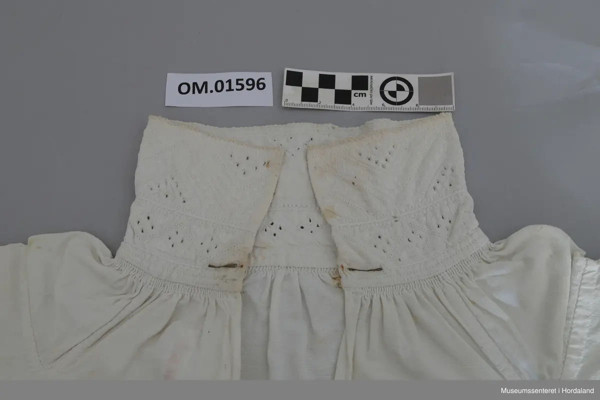 Kvit bomullskjorte med kvitsaumbroderi på krage. Kragen er høg og består av to border. To knapphol. Isett stoffbitar og rynking i hals. Splitt frå hals og ned halve skjorta. Kile under armane. Armane er rynka og har mansjettar utan broderi. Hekter. Ingen splitt i sidene. Handsauma. Eit hol i nakken. Flekkete.