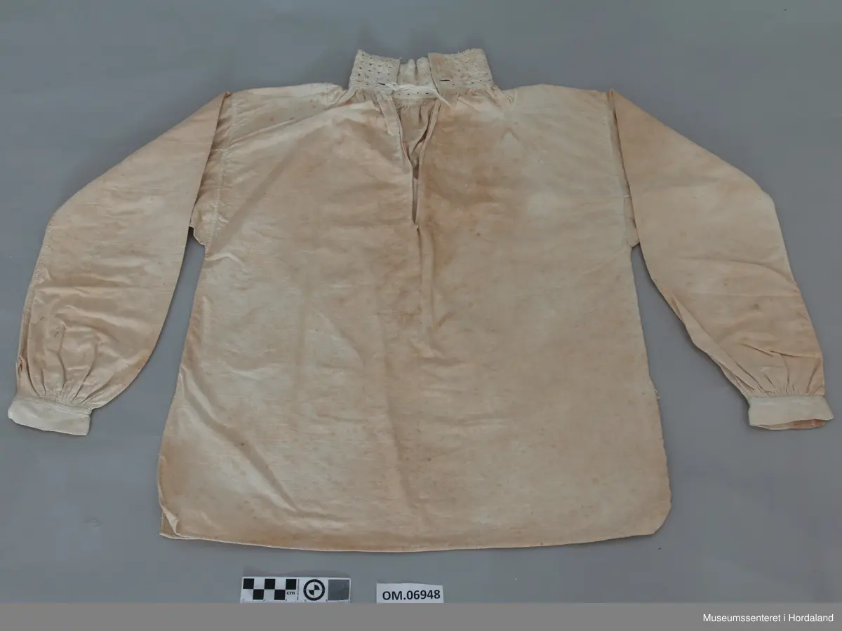 Kvit bomullskjorte med linmansjettar og krage. Kvitsaum på krage med firkantmotiv og holsaum. Rynka rundt hals og armar. Halssplitt. Ingen broderi på mansjettar. Kile under armane. Eine armen har isett stykke på langs. Skjorta har splittar i kvar side nede. Handsauma. Veldig flekkete og misfarga.