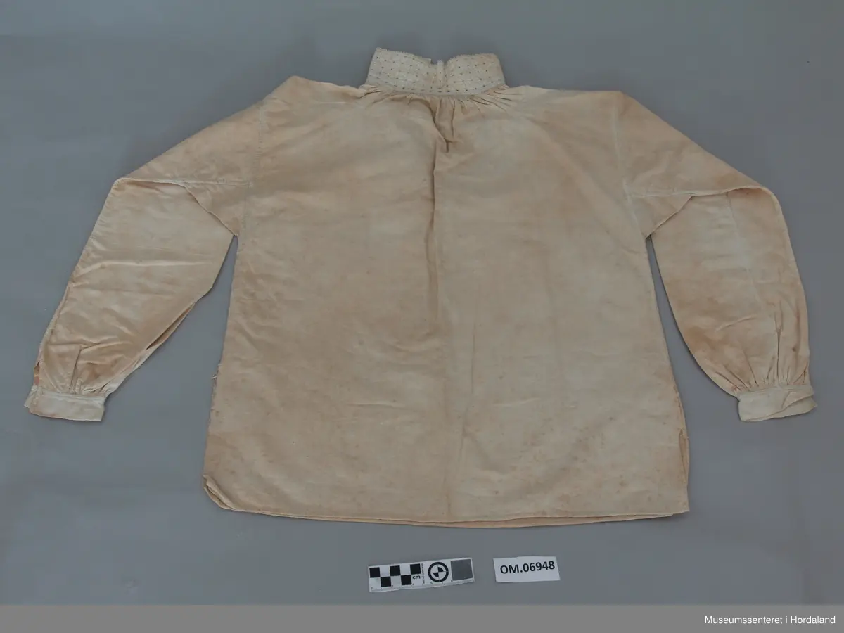 Kvit bomullskjorte med linmansjettar og krage. Kvitsaum på krage med firkantmotiv og holsaum. Rynka rundt hals og armar. Halssplitt. Ingen broderi på mansjettar. Kile under armane. Eine armen har isett stykke på langs. Skjorta har splittar i kvar side nede. Handsauma. Veldig flekkete og misfarga.