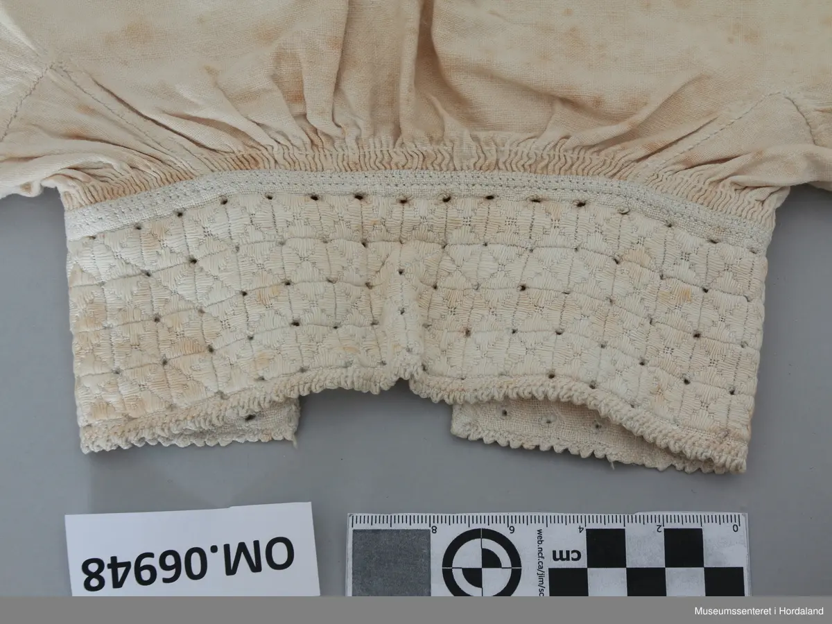Kvit bomullskjorte med linmansjettar og krage. Kvitsaum på krage med firkantmotiv og holsaum. Rynka rundt hals og armar. Halssplitt. Ingen broderi på mansjettar. Kile under armane. Eine armen har isett stykke på langs. Skjorta har splittar i kvar side nede. Handsauma. Veldig flekkete og misfarga.