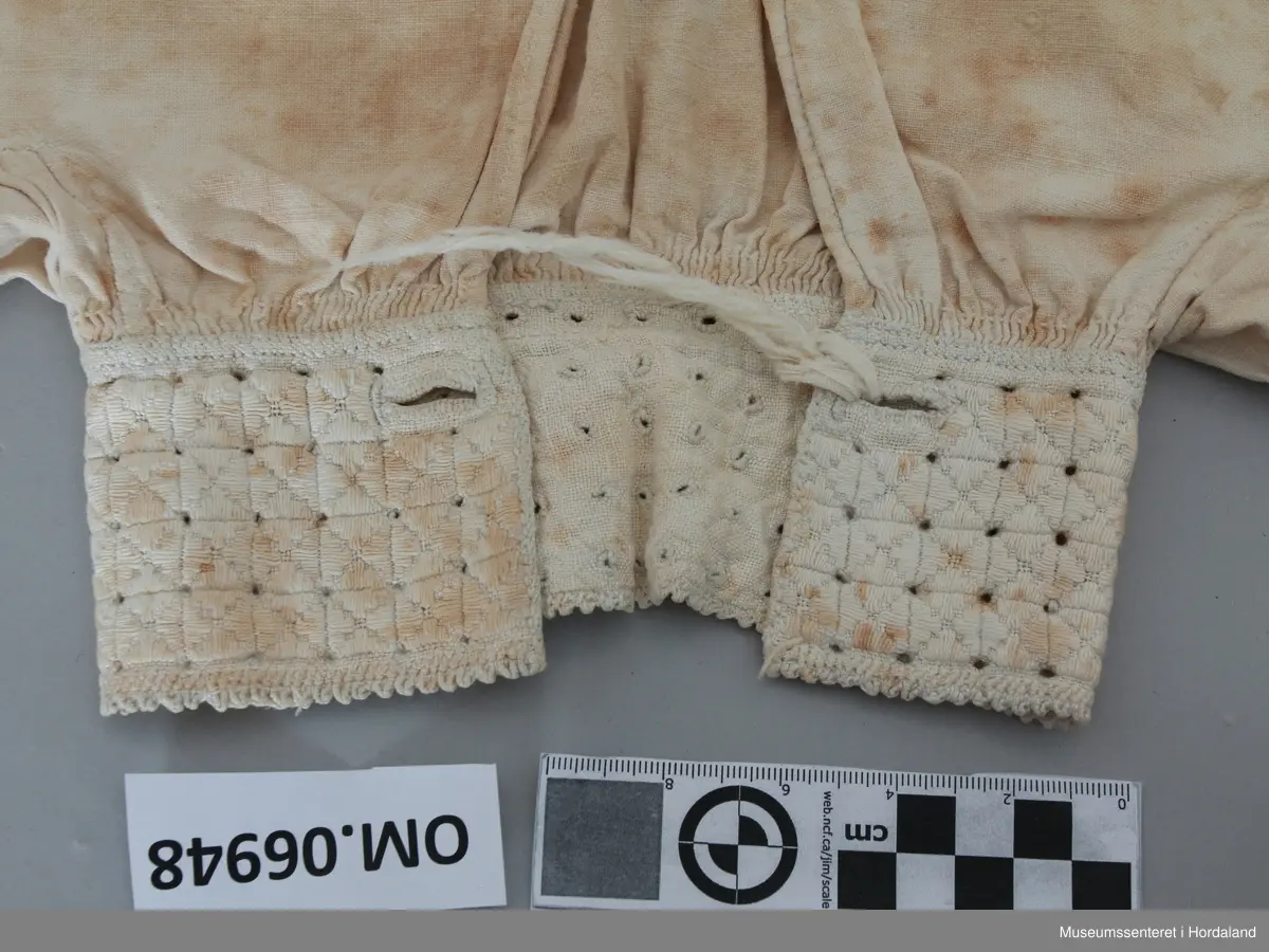 Kvit bomullskjorte med linmansjettar og krage. Kvitsaum på krage med firkantmotiv og holsaum. Rynka rundt hals og armar. Halssplitt. Ingen broderi på mansjettar. Kile under armane. Eine armen har isett stykke på langs. Skjorta har splittar i kvar side nede. Handsauma. Veldig flekkete og misfarga.