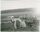 Studenter, kvinnor och män sittande vid bord ute på gräs, skog i bakgrunden, sannolikt Uppland omkring 1900