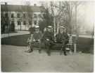 Tre män iklädda kostym och studentmössor sittande på bänk i Universitetsparken, Uppsala omkring 1900