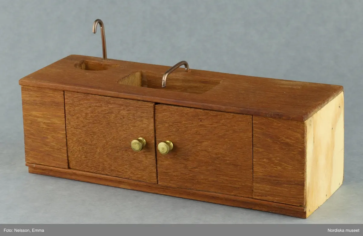 Inventering Sesam 1996-1999:
B 77,5, Djup 36,0, H 119,0 cm
Dockskåp av furu med mellanväggar av plywood, målat i gräddgult, fasaden "putsad" med färg blandad med sågspån. Dockskåpet har dubbeldörrar med lås och en fasad på gångjärn med en dörr och 10 fönster, inspirerad av det första kollektivhuset i funkisstil på John Erikssons gata. Dockskåpet är befolkat av fam. Björnerholm. Gunvor Svartz-Malmberg f. 1925-6 var ca 10 år när hon fick skåpet. 
LS 85
Helena Carlsson
