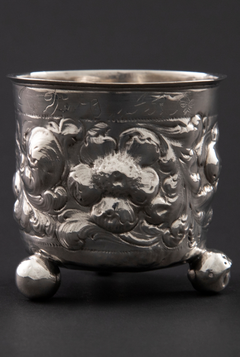 Sølvbeger med svakt konisk form som hviler på tre kuleføtter. Korpus er dekorert med rikt drevet blomsterdekor.