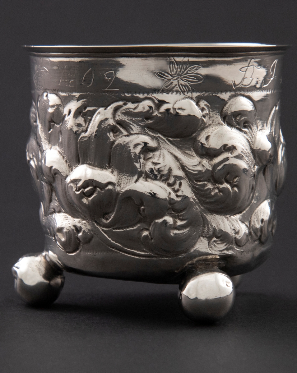Sølvbeger med svakt konisk form som hviler på tre kuleføtter. Korpus er dekorert med rikt drevet blomsterdekor.