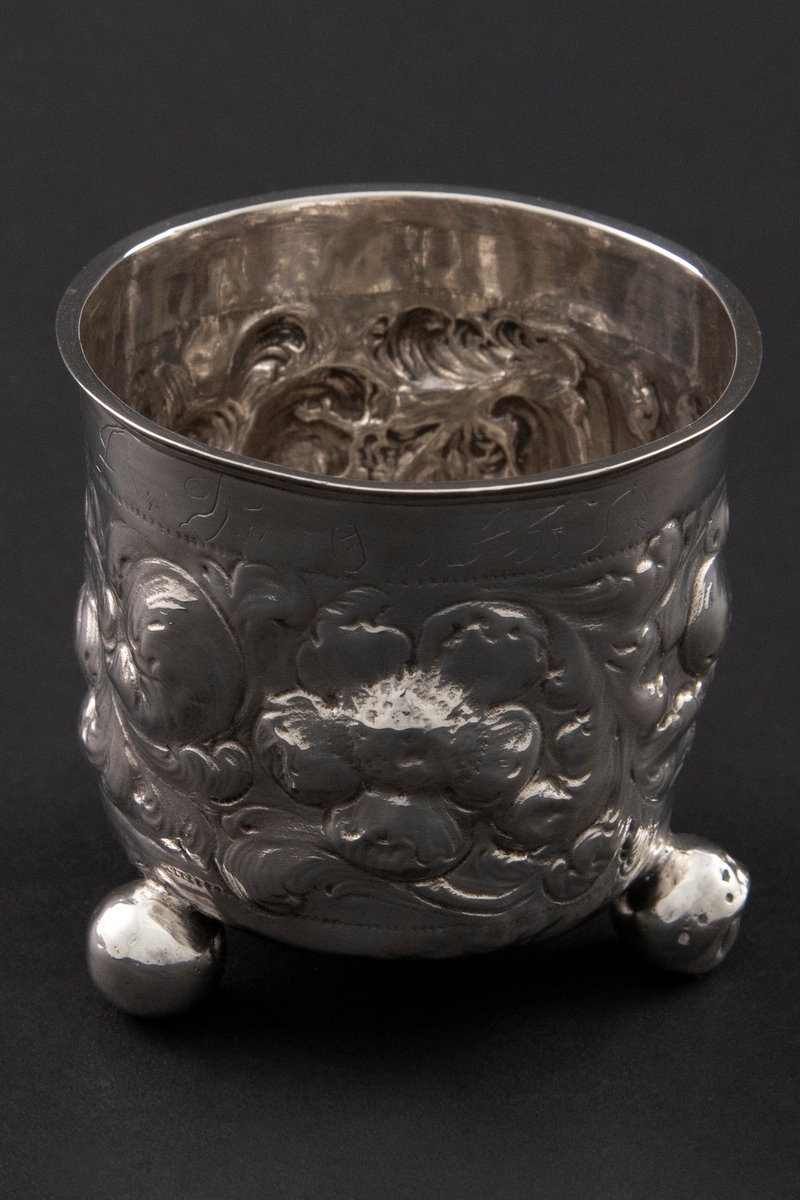 Sølvbeger med svakt konisk form som hviler på tre kuleføtter. Korpus er dekorert med rikt drevet blomsterdekor.