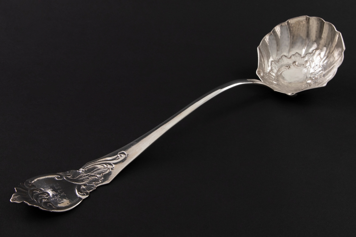 Suppeøse av sølv med muslingformet blad med drevet rocaillemotiv i bunn. Skaftet er S-formet og ender i en kartusj omringet av asymmetriske rocailleornamenter.
