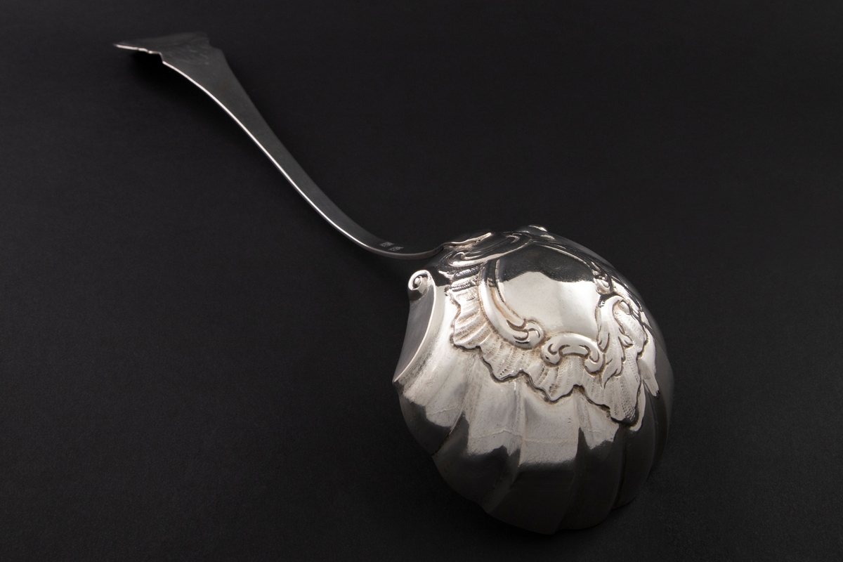 Suppeøse av sølv med muslingformet blad med drevet rocaillemotiv i bunn. Skaftet er S-formet og ender i en kartusj omringet av asymmetriske rocailleornamenter.