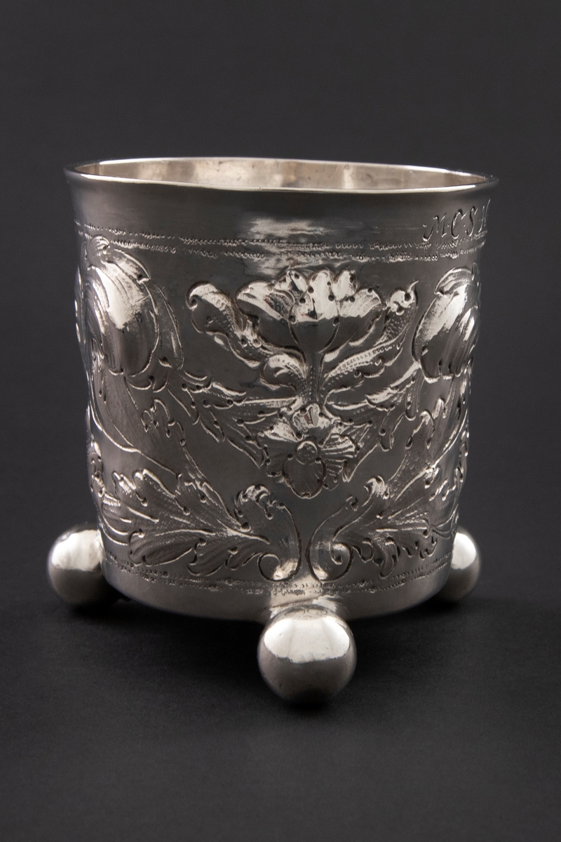 Sølvbeger med svakt konisk form som hviler på tre kuleføtter. Korpus er dekorert med drevet, siselert og gravert blomsterornamentikk.