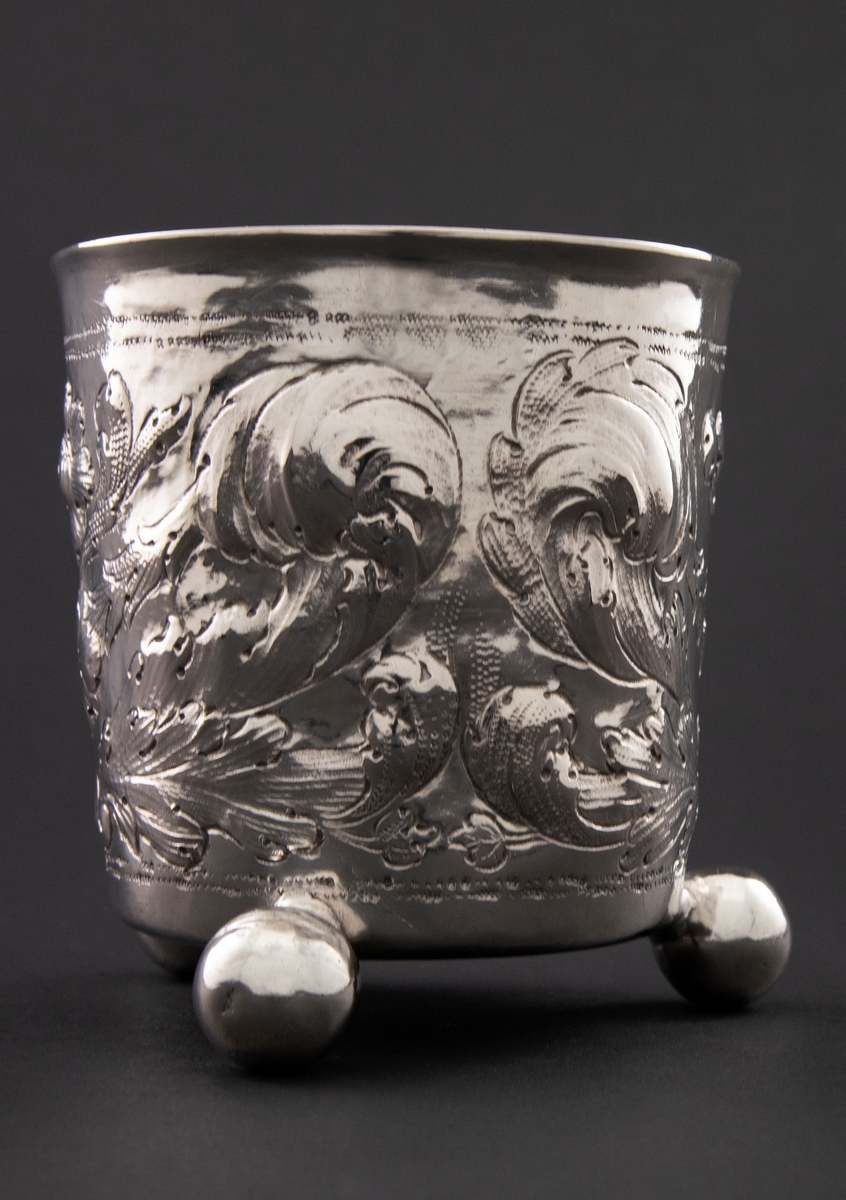 Sølvbeger med svakt konisk form som hviler på tre kuleføtter. Korpus er dekorert med drevet, siselert og gravert blomsterornamentikk.