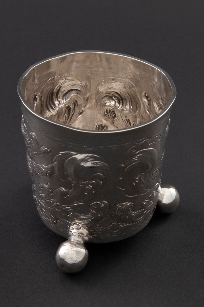 Sølvbeger med svakt konisk form som hviler på tre kuleføtter. Korpus er dekorert med drevet, siselert og gravert blomsterornamentikk.
