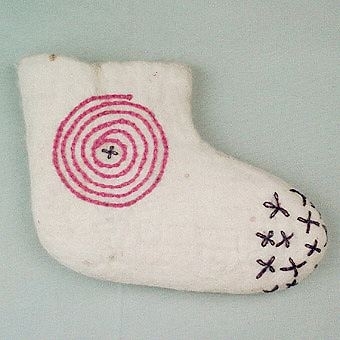 Socka tovad av naturvit ull, troligtvis gobelinull. Sockan har en naturlig avslutad rak kant. Broderi på skaftets utsida med rosa tvåtrådigt ullgarn i form av en spiral i kedjesöm och i mitten en lila blomma i öglesöm. På sockans spets är broderat lila blommor i öglesöm. Högersocka.