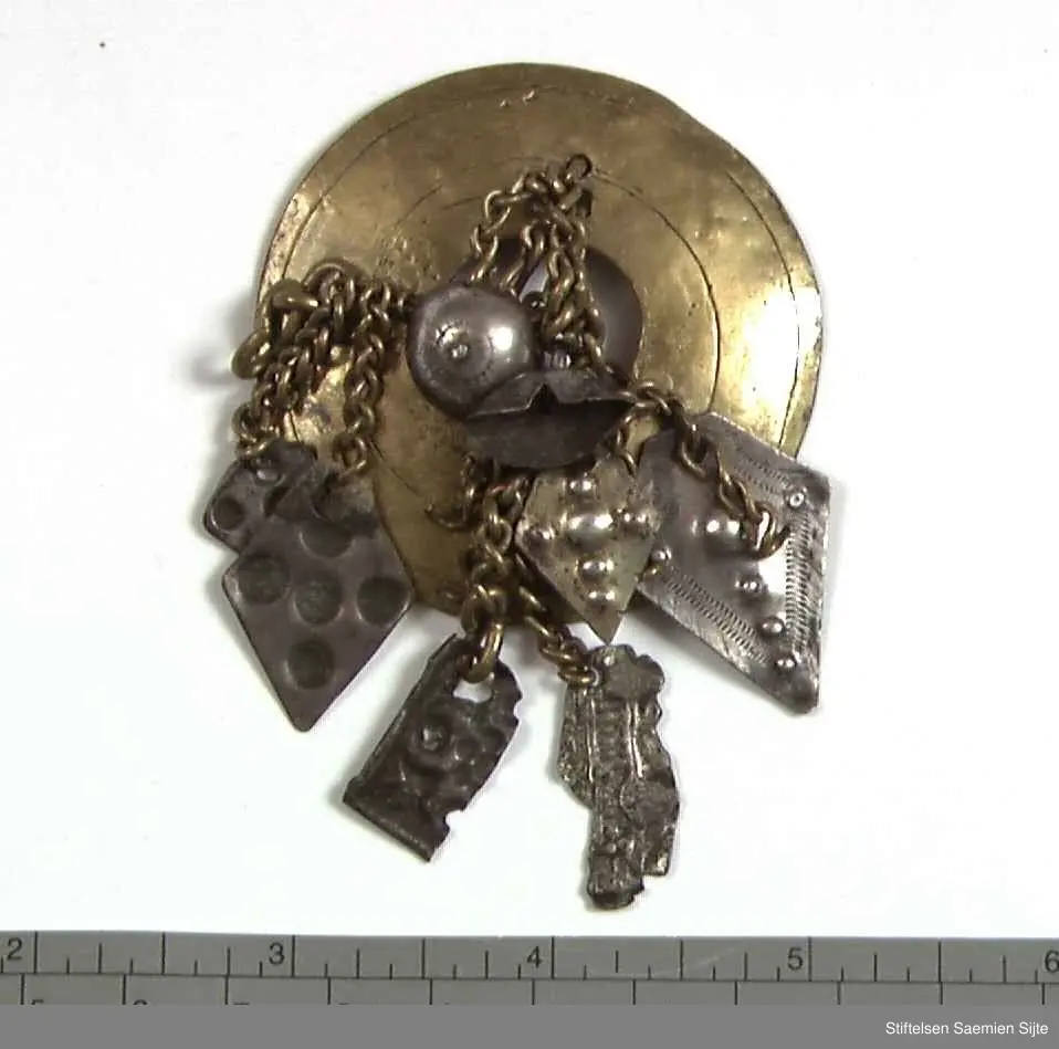 Rund, svakt konveks messingplate med hull i midten. Fastmonterte messinglenker med dekorative 8 sølvsmykker i endene som er punslet.