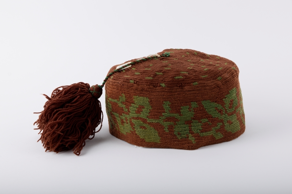 Kalott med stor dusk. Brun med grønt mønster. Rester av svart tekstil innvendig langs randen, dermed har luen trolig vært fôret.