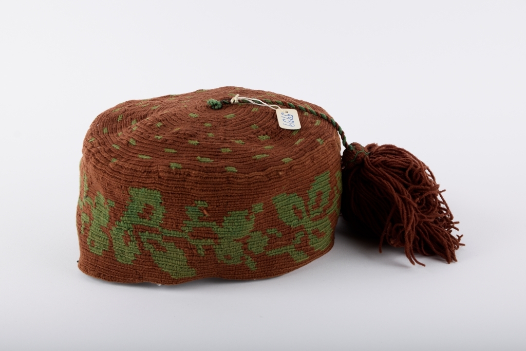 Kalott med stor dusk. Brun med grønt mønster. Rester av svart tekstil innvendig langs randen, dermed har luen trolig vært fôret.