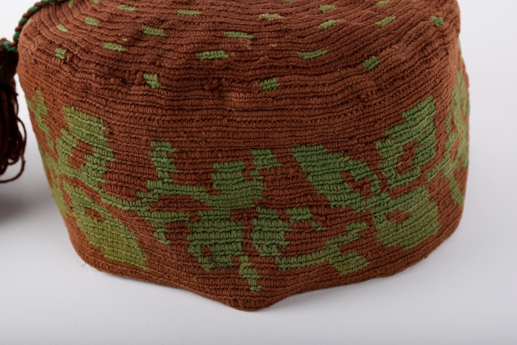 Kalott med stor dusk. Brun med grønt mønster. Rester av svart tekstil innvendig langs randen, dermed har luen trolig vært fôret.