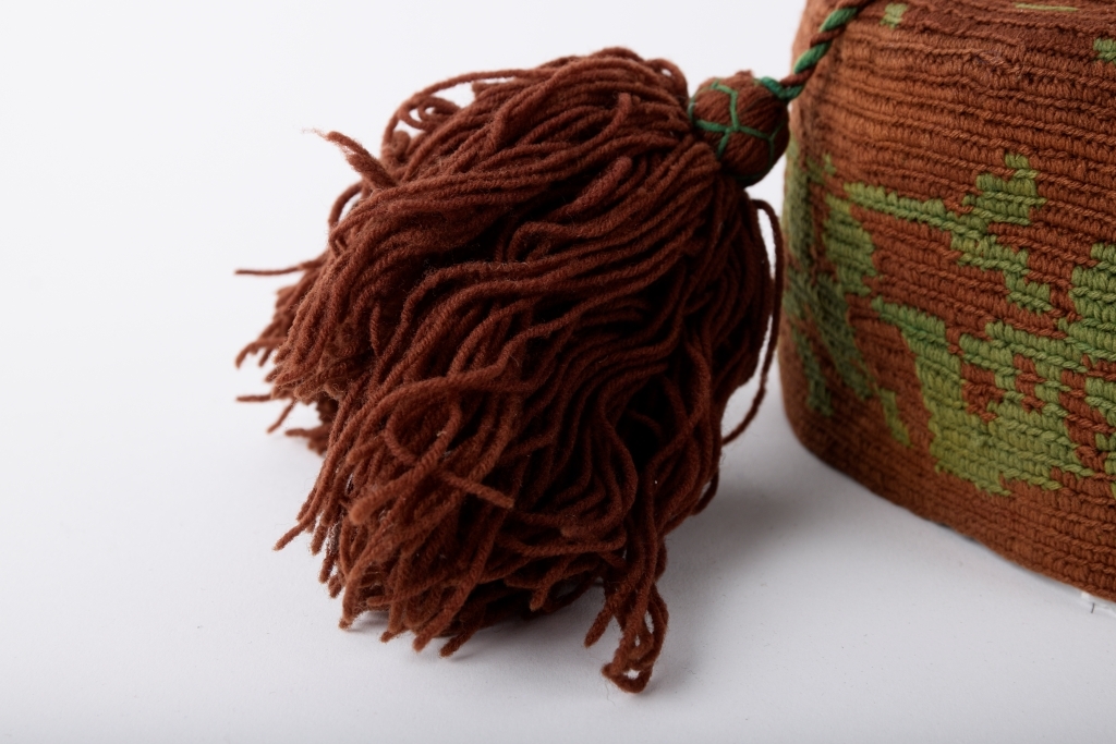 Kalott med stor dusk. Brun med grønt mønster. Rester av svart tekstil innvendig langs randen, dermed har luen trolig vært fôret.