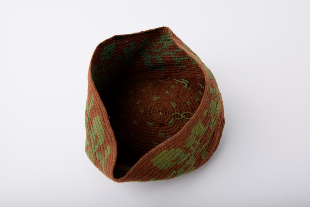 Kalott med stor dusk. Brun med grønt mønster. Rester av svart tekstil innvendig langs randen, dermed har luen trolig vært fôret.