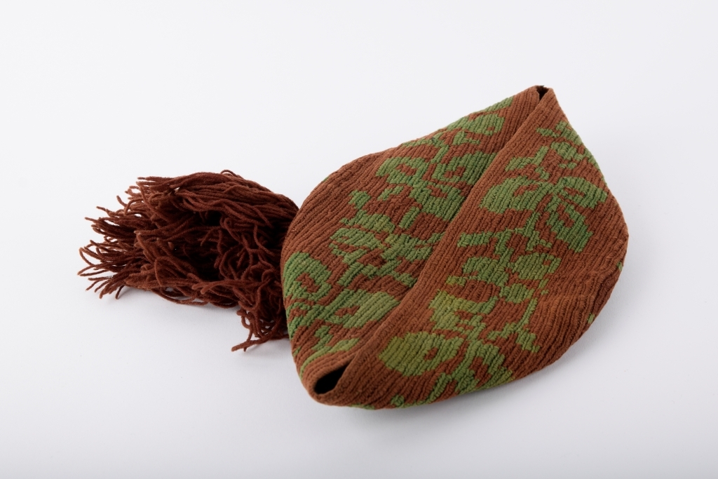 Kalott med stor dusk. Brun med grønt mønster. Rester av svart tekstil innvendig langs randen, dermed har luen trolig vært fôret.