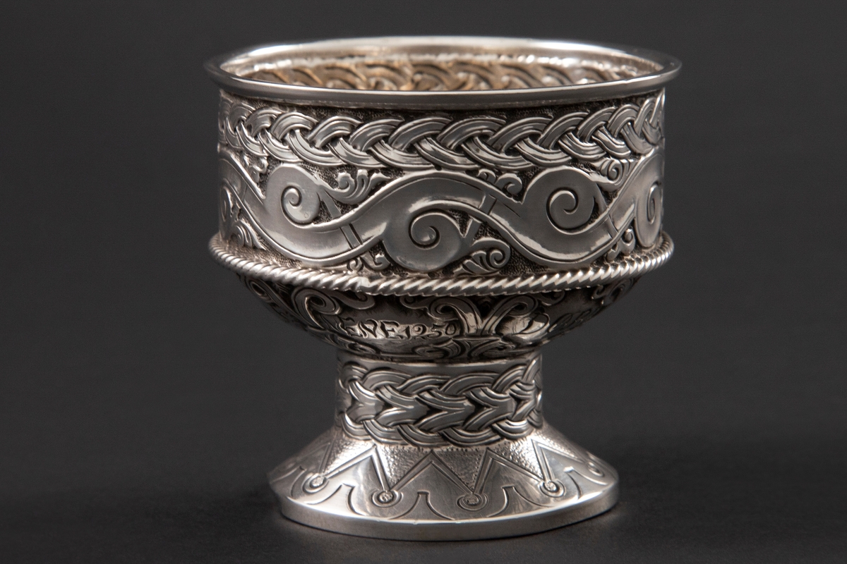 Beger i sølv med sirkulær form. Foten er dekorert med geometriske mønstre, stetten med flettemønster og kupa med bølge- og flettemønster. Rundt kupa er også et pålagt snodd bånd.