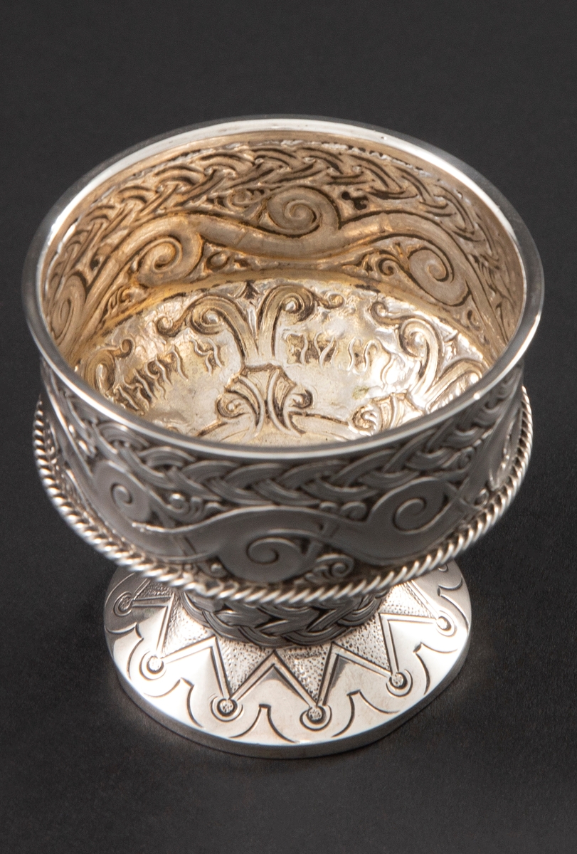 Beger i sølv med sirkulær form. Foten er dekorert med geometriske mønstre, stetten med flettemønster og kupa med bølge- og flettemønster. Rundt kupa er også et pålagt snodd bånd.