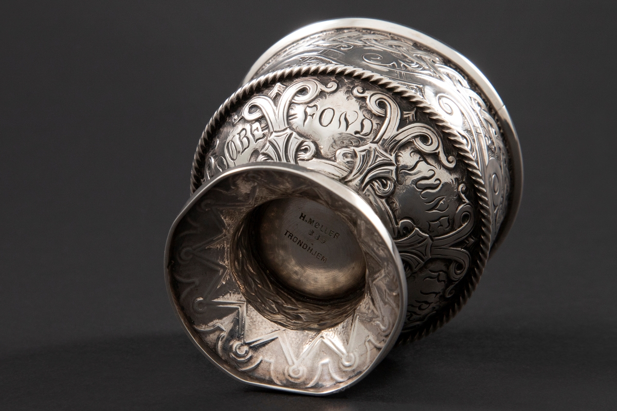 Beger i sølv med sirkulær form. Foten er dekorert med geometriske mønstre, stetten med flettemønster og kupa med bølge- og flettemønster. Rundt kupa er også et pålagt snodd bånd.