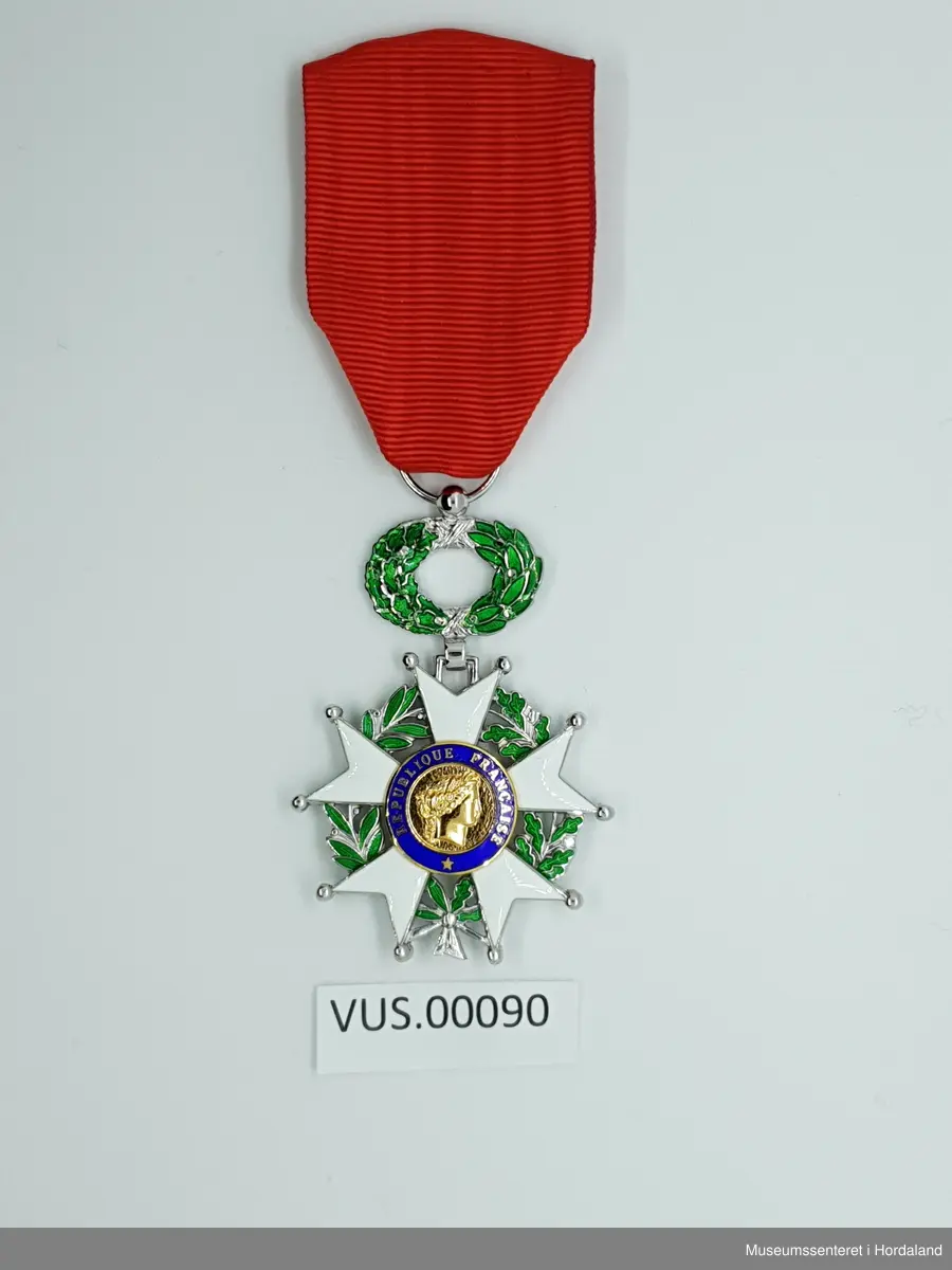 Hvit kort på grønne laurbærkranser. Senter i korste med kvinnerprofil i gull in blå sirkel. Kvinnen symboliserer den fransk republikken. Tekst skrevet i den blå sirkenen rundt senter. 

