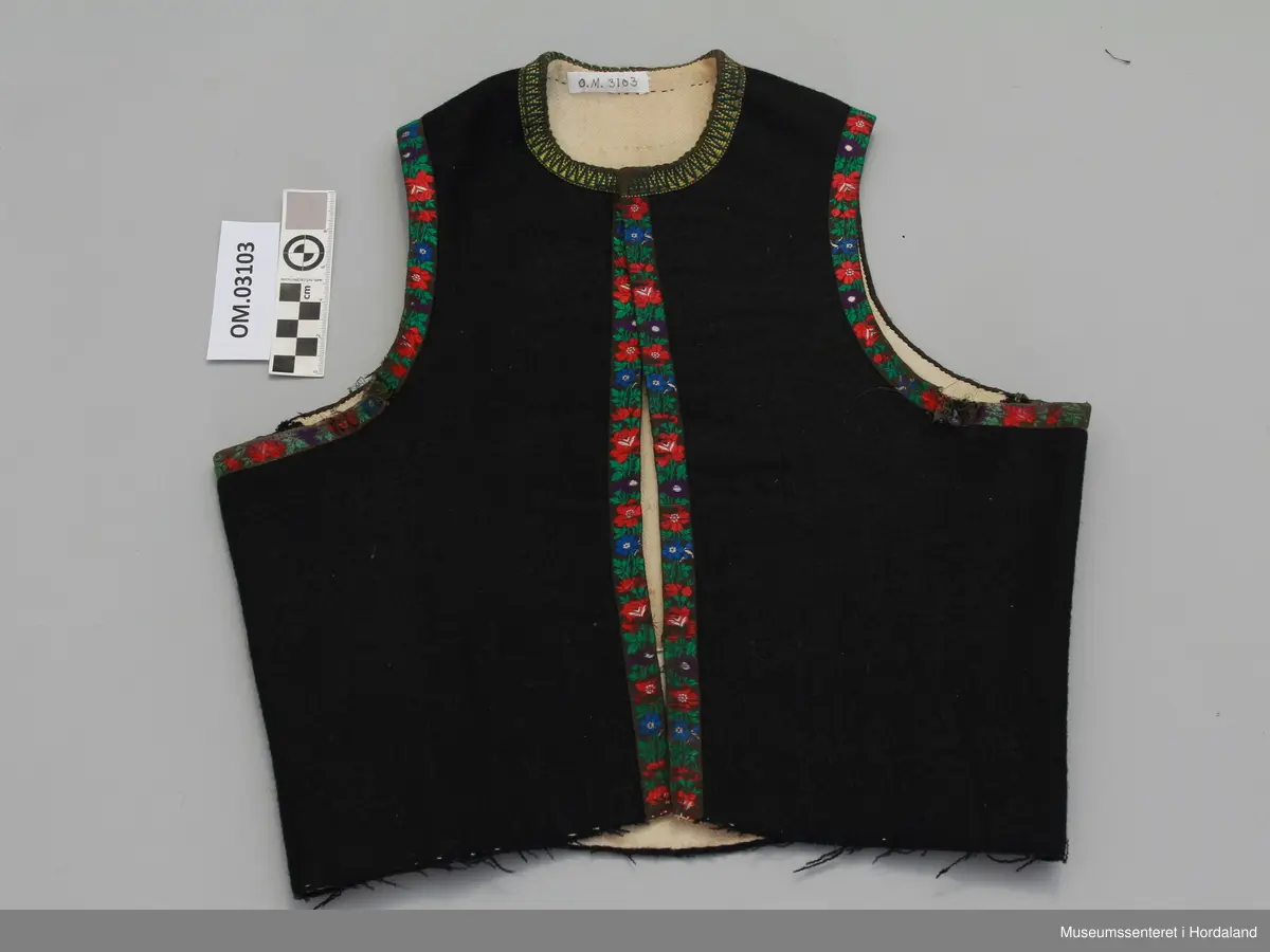 Svart liv i ull med bomullsfôr. Roseband av silke i frontopninga og rundt armholene, gul/grøn kvardekant i silke rundt hals.