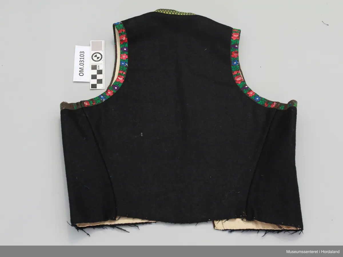 Svart liv i ull med bomullsfôr. Roseband av silke i frontopninga og rundt armholene, gul/grøn kvardekant i silke rundt hals.