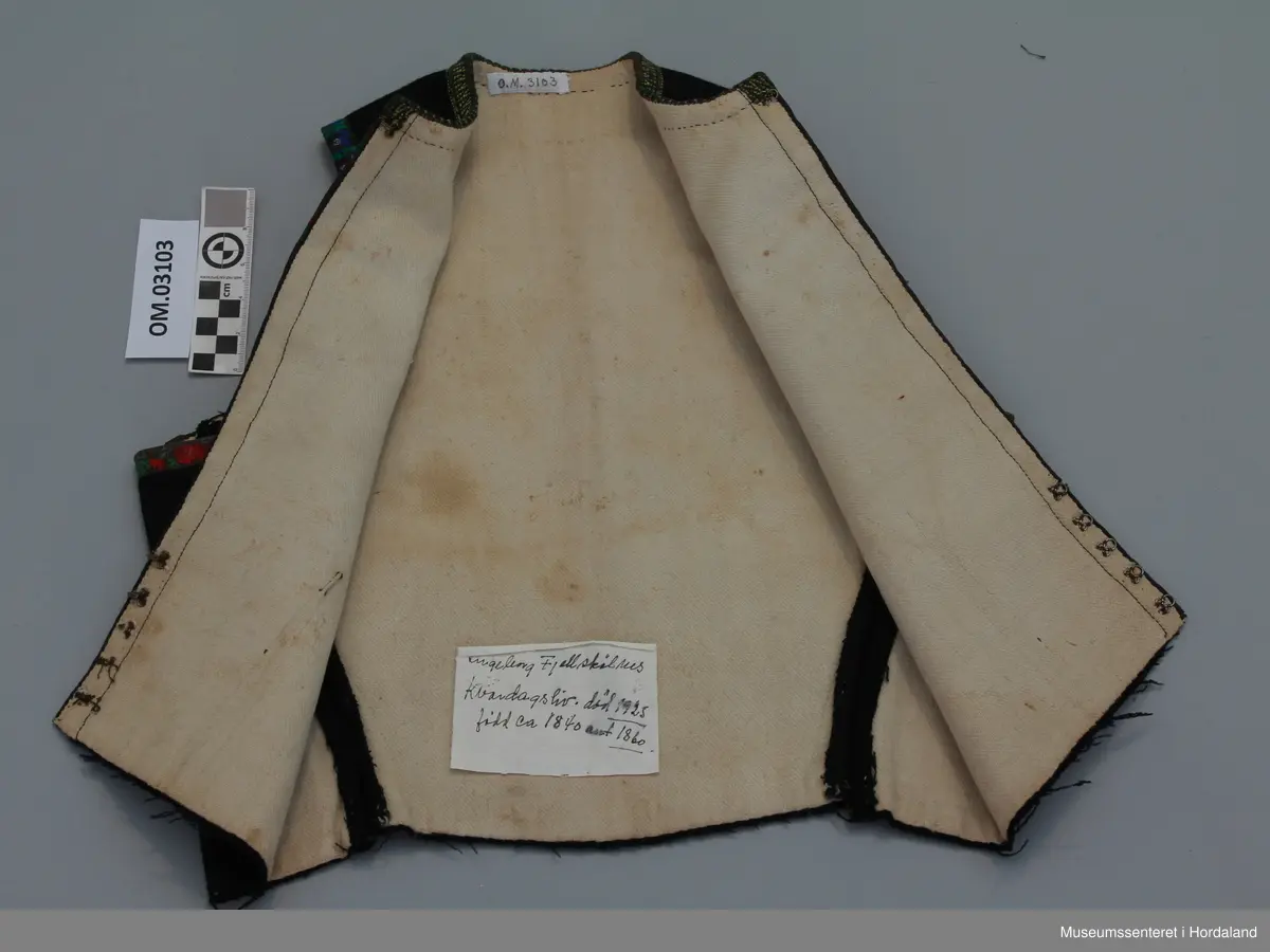 Svart liv i ull med bomullsfôr. Roseband av silke i frontopninga og rundt armholene, gul/grøn kvardekant i silke rundt hals.