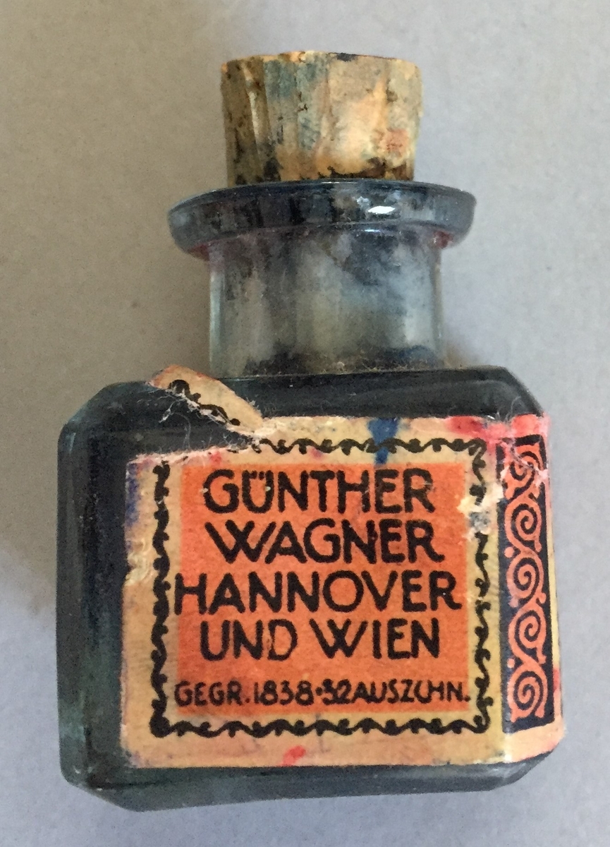 Lite blekkhus med kork. Korken er knekt, Sjølve blekkhuset er i glas og har raud etikett med teksten"Gunter wagner Hannover und Wien Rote Metalltinte Schutzmarks Pelikan"