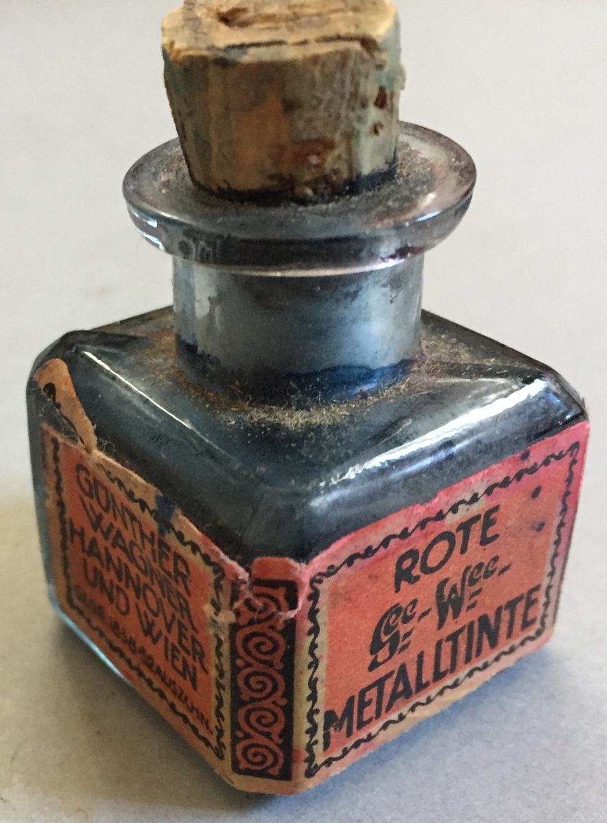 Lite blekkhus med kork. Korken er knekt, Sjølve blekkhuset er i glas og har raud etikett med teksten"Gunter wagner Hannover und Wien Rote Metalltinte Schutzmarks Pelikan"