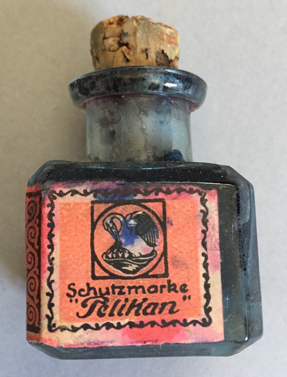 Lite blekkhus med kork. Korken er knekt, Sjølve blekkhuset er i glas og har raud etikett med teksten"Gunter wagner Hannover und Wien Rote Metalltinte Schutzmarks Pelikan"