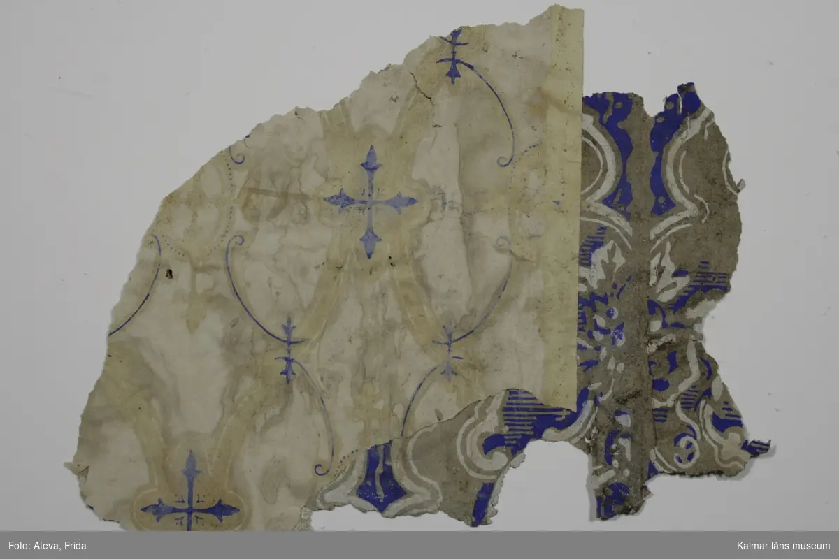 KLM 43762:13:1. Tapet av papper. 5 st tapetbitar. Tryckt tapet med vit botten och utsparat mönster som bildar medaljonger. Ultramarinblåa detaljer. Närmast underliggande lager har tryck i ultramarinblått och vitt på ofärgad botten, med datering till 1840-1850-tal. På en av bitarna sitter ett lager med KLM 43762:13:2, detta blir då det understa lagret.