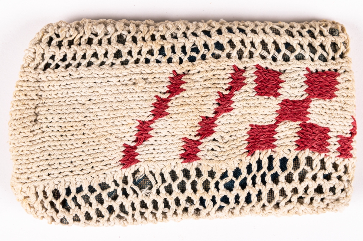 Dyna, till docka.
Stickad remsa med röda rutor. Hopsydd med flätsöm. Fylld innerdyna.
Mönsterstickad med vitt och rött bomullsgarn. Blåvitt bomullstyg och fyllning.