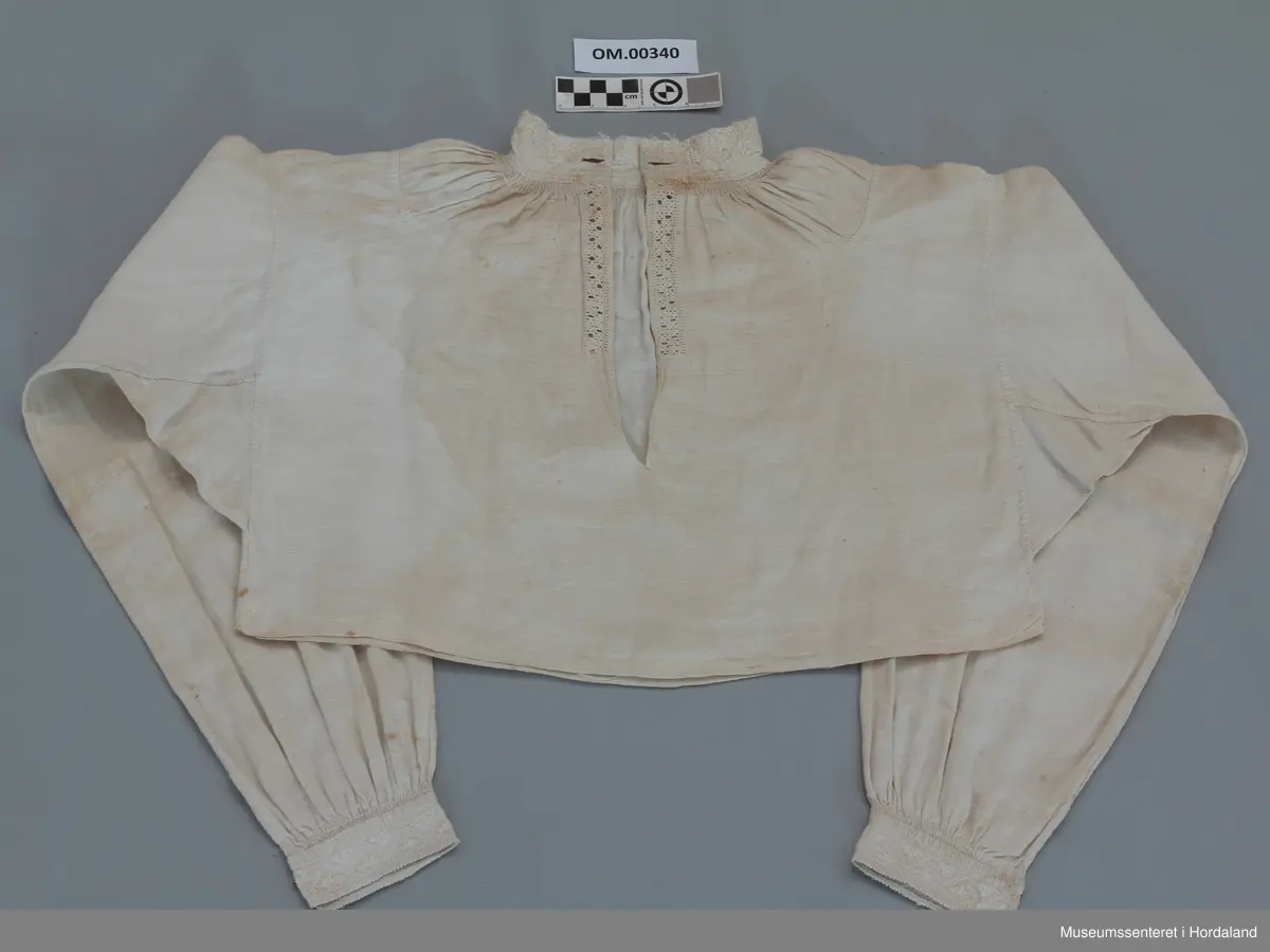 Kvit, kort linskjorte der krage og armmansjettar er sydd med kvitsaum. Stolpen i bringa er sydd med enkel uttrekkssaum. Kilar under armane og oppå skuldrane. Rynker rundt hals og framme på armane. Kragen har eit knapphol på kvar side. Sliten øvrekant på kragen.