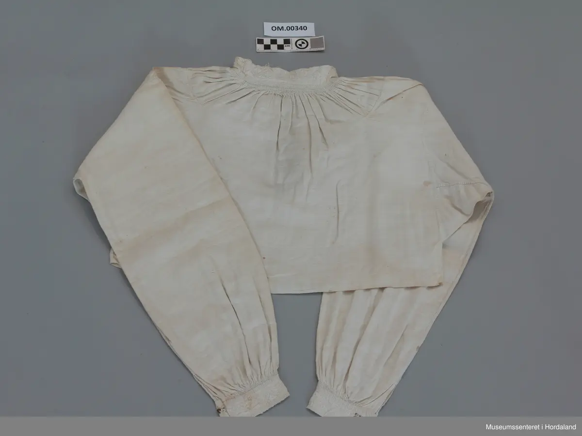 Kvit, kort linskjorte der krage og armmansjettar er sydd med kvitsaum. Stolpen i bringa er sydd med enkel uttrekkssaum. Kilar under armane og oppå skuldrane. Rynker rundt hals og framme på armane. Kragen har eit knapphol på kvar side. Sliten øvrekant på kragen.