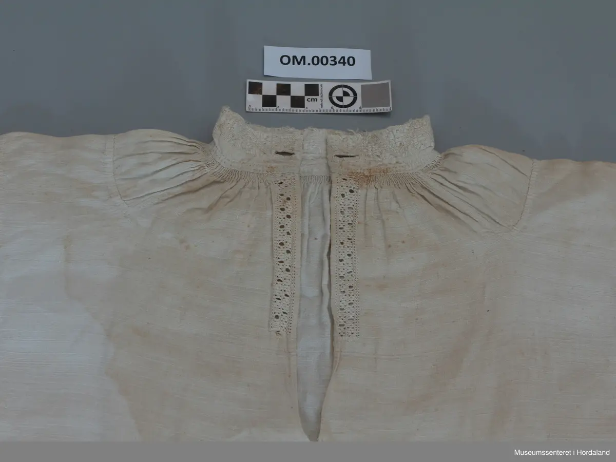 Kvit, kort linskjorte der krage og armmansjettar er sydd med kvitsaum. Stolpen i bringa er sydd med enkel uttrekkssaum. Kilar under armane og oppå skuldrane. Rynker rundt hals og framme på armane. Kragen har eit knapphol på kvar side. Sliten øvrekant på kragen.